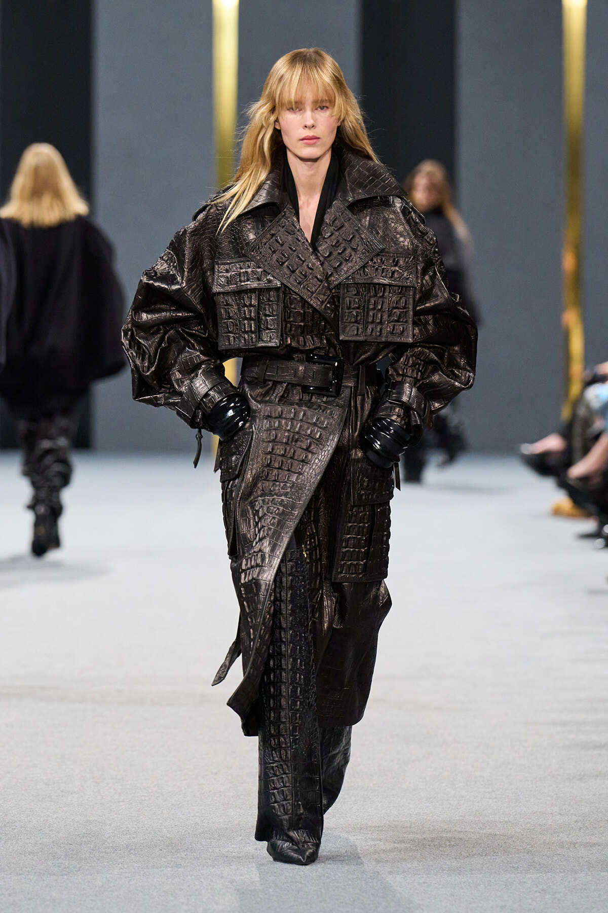 Female model on a runway wearing a large, dark crocodile-embossed leather trench with a wide belt and gloves.