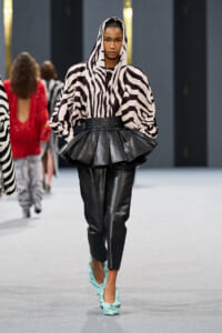 Model walking a runway in a zebra-print hooded top with oversized sleeves, a black peplum belt, and black leather pants with turquoise heels.