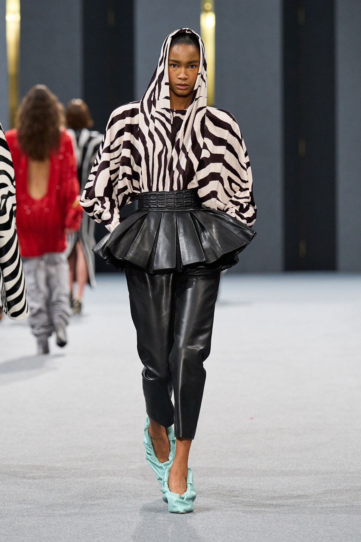 Model walking a runway in a zebra-print hooded top with oversized sleeves, a black peplum belt, and black leather pants with turquoise heels.
