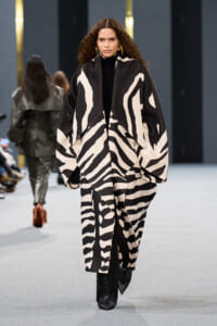Runway model walking in a bold black-and-white zebra-patterned oversized coat over a black turtleneck, with black boots and gold earrings.
