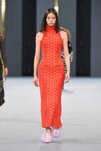 Model walking the runway in a sleeveless red woven knit maxi dress with a high collar and lavender fluffy slippers.