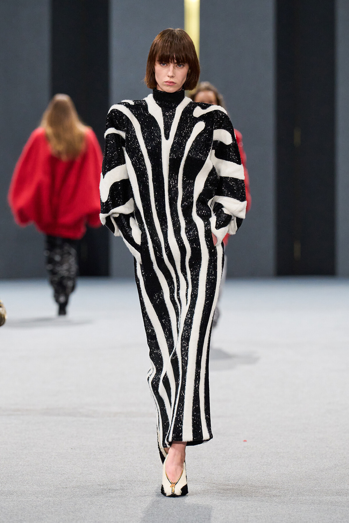 Model walking the runway in a bold black-and-white zebra-stripe knit jumpsuit with voluminous sleeves and a high neckline.