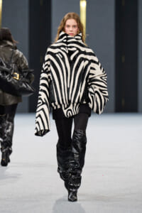 Model walking a runway in a black-and-white zebra-print oversized coat with a high collar, paired with black leggings and slouchy knee-high boots.