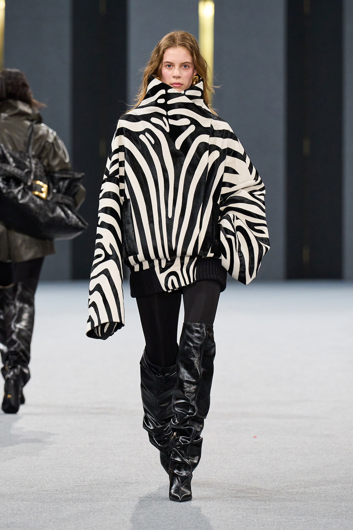 Model walking a runway in a black-and-white zebra-print oversized coat with a high collar, paired with black leggings and slouchy knee-high boots.