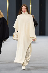 Model walks the runway in a cream oversized ribbed knit poncho with a high turtleneck, paired with matching boots and a beige handbag.