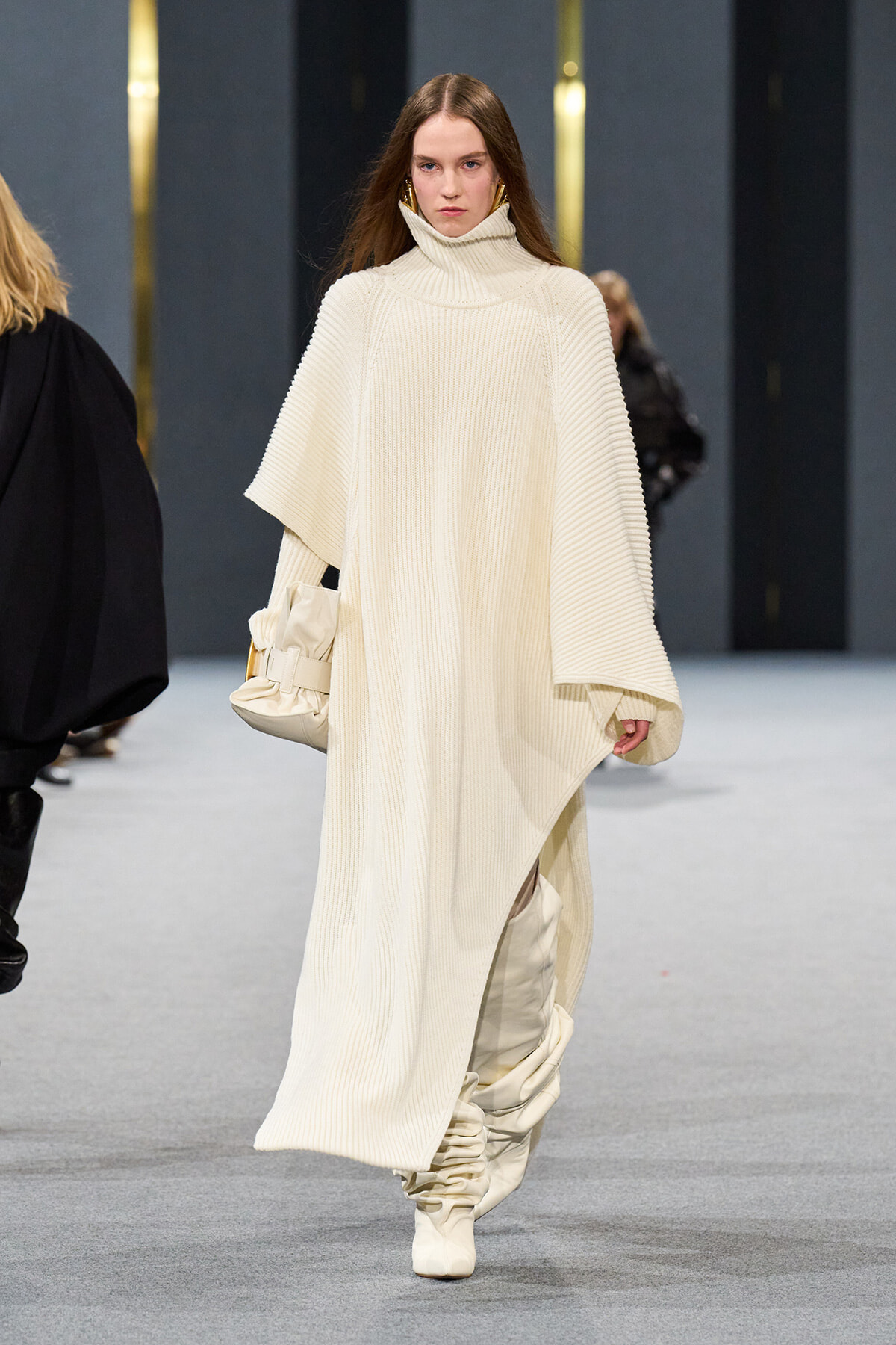 Model walks the runway in a cream oversized ribbed knit poncho with a high turtleneck, paired with matching boots and a beige handbag.