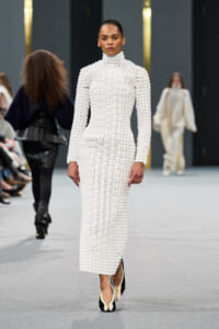 Model on a runway wearing a white high-neck, square-block knit dress with matching shoes.