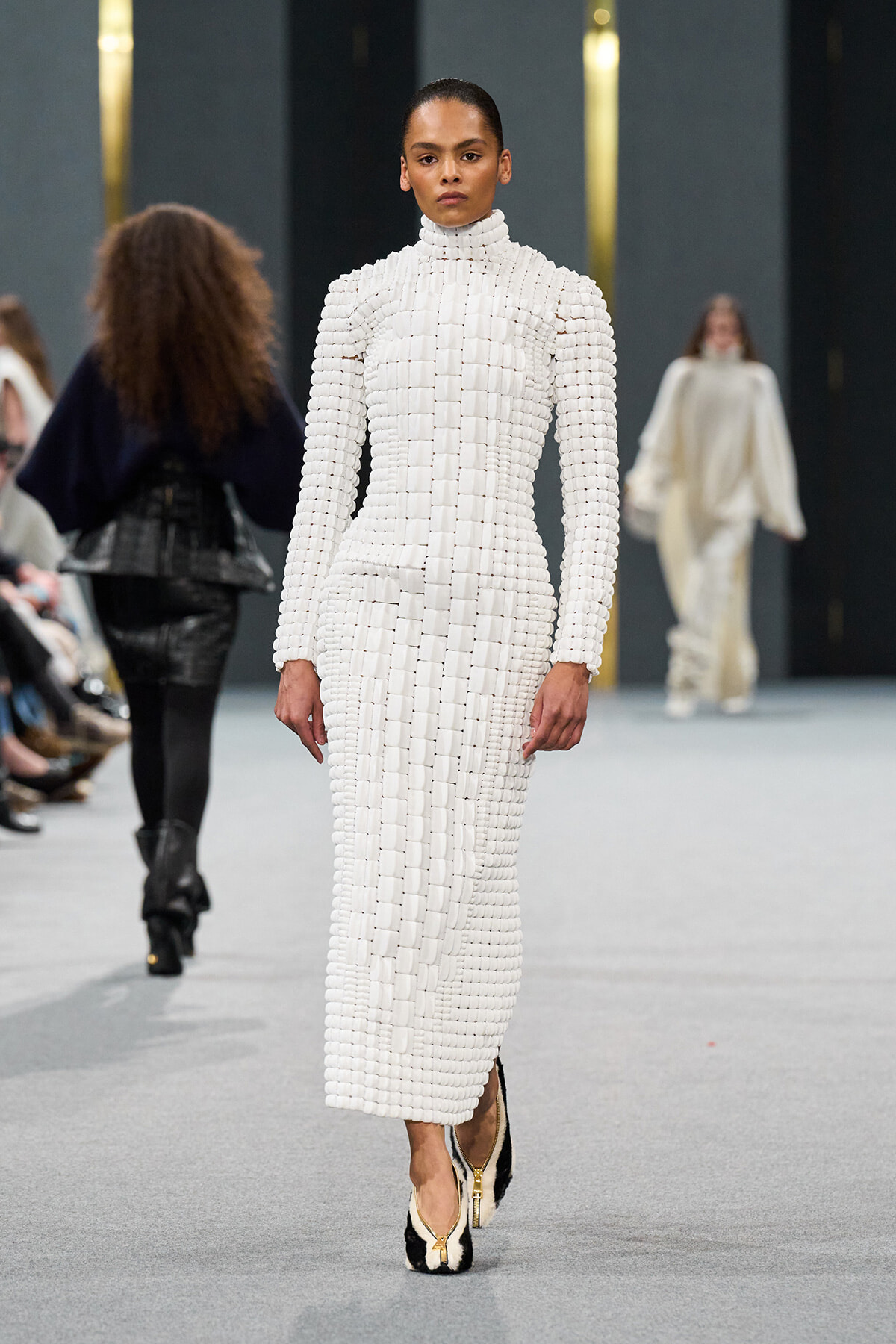 Model on a runway wearing a white high-neck, square-block knit dress with matching shoes.