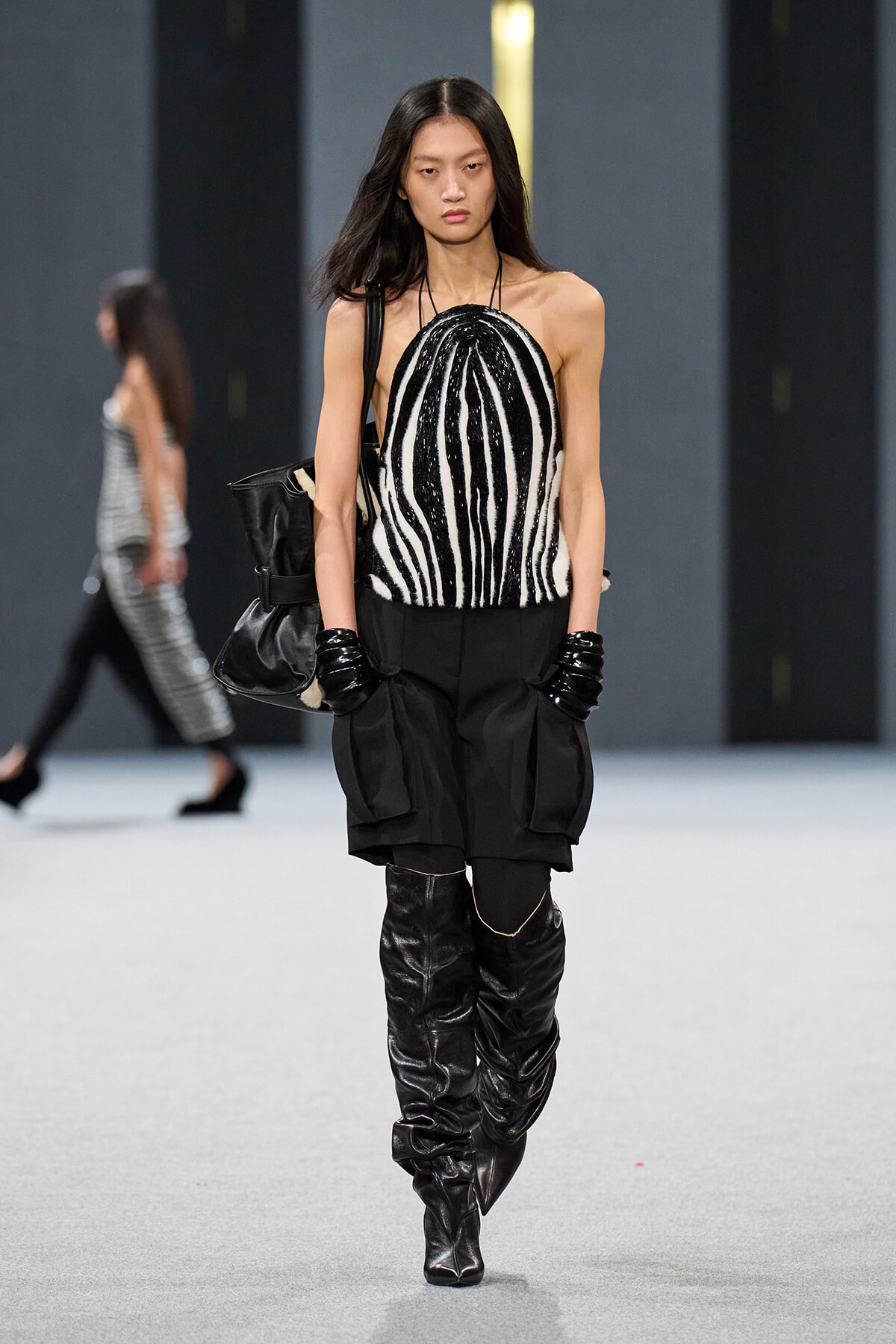 Model on a fashion runway wearing a black-and-white zebra-print halter top with black oversized shorts, gloves, and tall black boots, carrying a black bag.