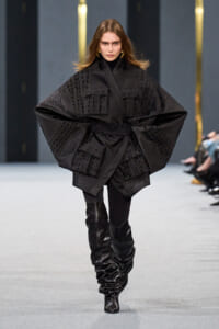 Model walks the runway in an oversized black textured cape with wide sleeves and a belt, paired with black leggings and slouchy knee-high boots.