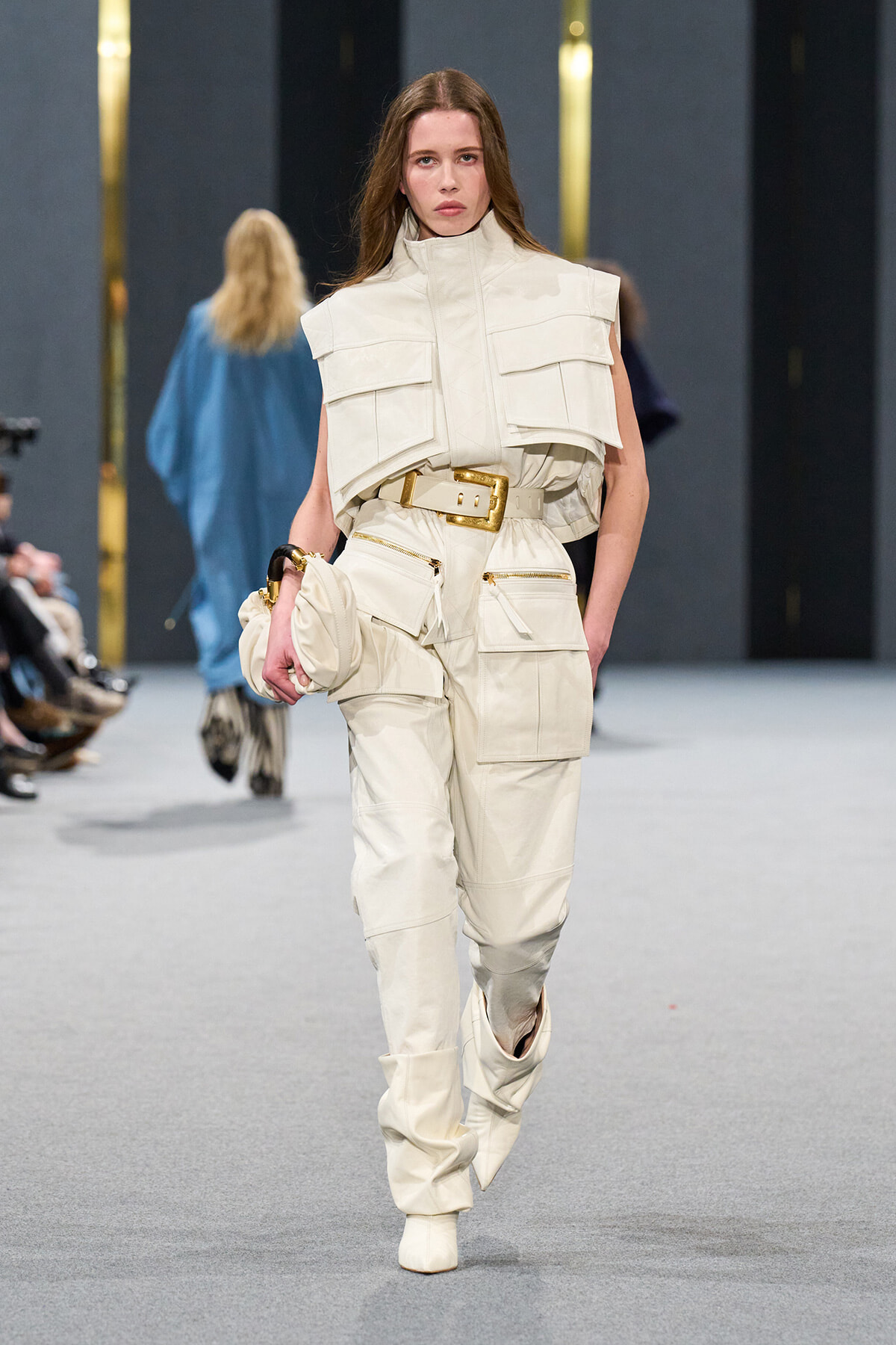 Model walks the runway in an off-white utility jumpsuit with multiple large pockets and a wide gold belt, high collar and matching boots.