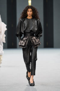 Model walks the runway in a glossy black crocodile-textured ensemble with oversized puff sleeves and a structured peplum belt over matching tailored pants.