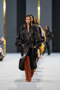 Model walks the runway in a black belted leather trench coat with brown fur boots; other models follow in the line behind.