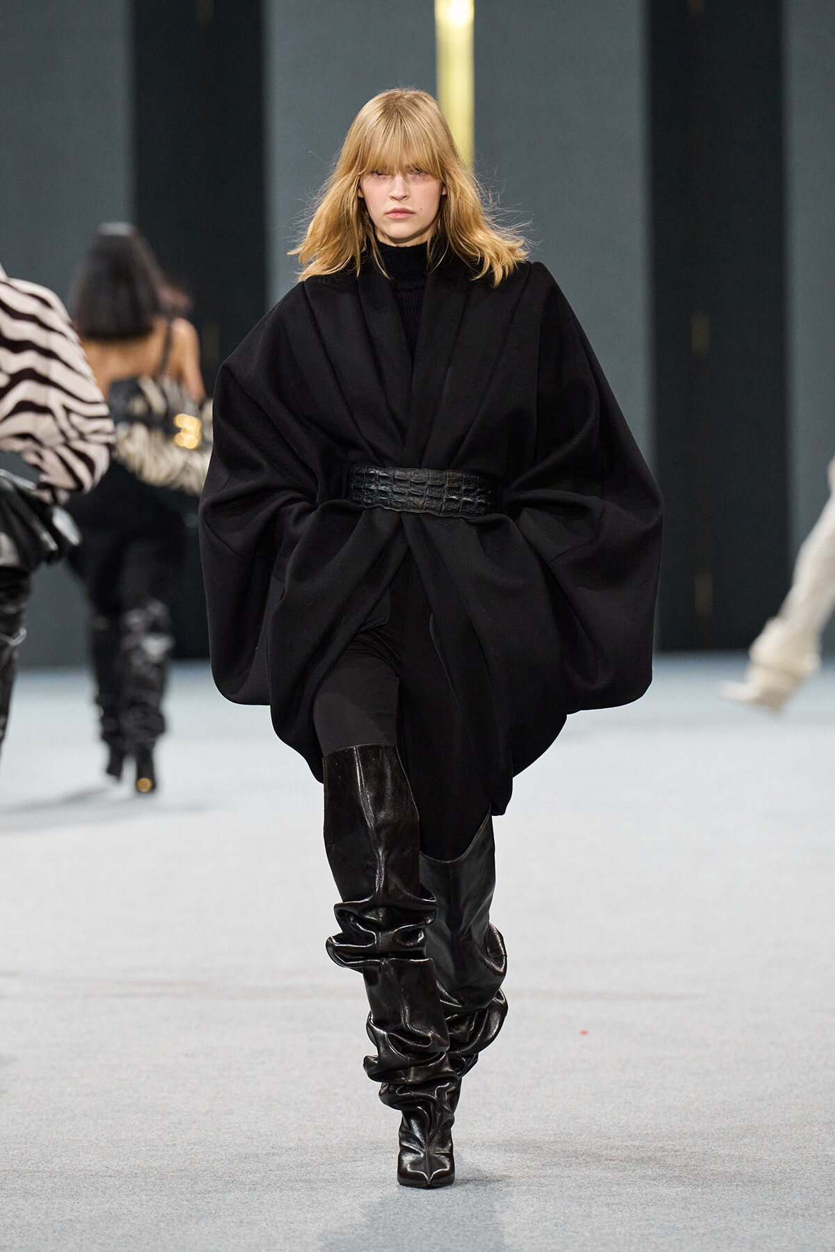Model walking a runway in a voluminous black cape dress cinched with a wide textured belt, paired with knee-high shiny black boots.