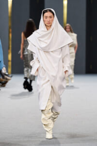 Model walking a runway in an all-white, draped ensemble with oversized scarf-like hood and slouchy boots.