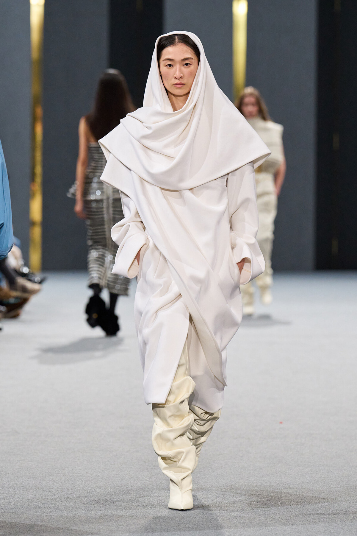 Model walking a runway in an all-white, draped ensemble with oversized scarf-like hood and slouchy boots.