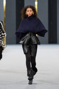 Model walking a runway in an oversized navy sweater with a black leather peplum waist, textured shorts, black tights, and slouchy black boots.