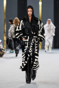 Model walking a fashion show in a black-and-white oversized patterned coat with a hood and glossy black boots.