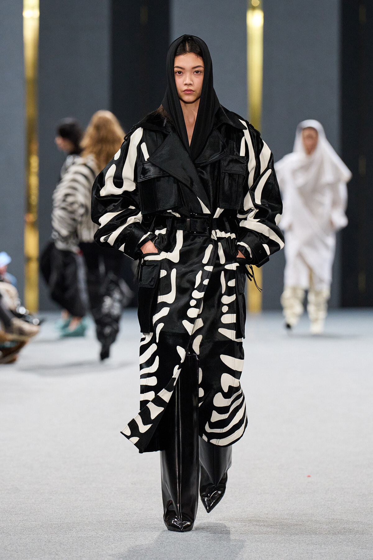 Model walking a fashion show in a black-and-white oversized patterned coat with a hood and glossy black boots.