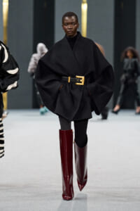 Model walking a fashion runway in an oversized black cape with a wide gold-buckle belt, black turtleneck, black leggings, and glossy burgundy knee-high boots.