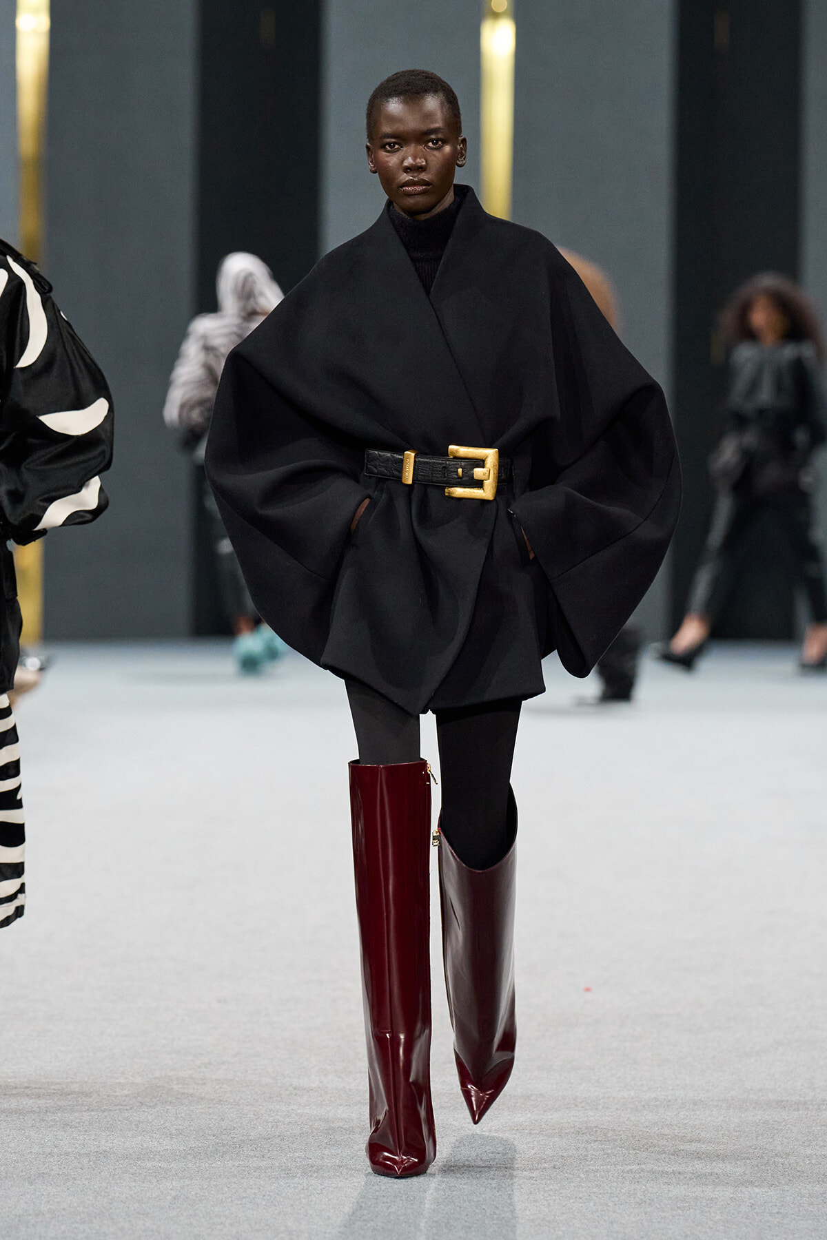 Model walking a fashion runway in an oversized black cape with a wide gold-buckle belt, black turtleneck, black leggings, and glossy burgundy knee-high boots.
