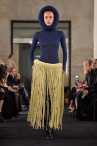 Runway model in a navy hooded bodysuit with a cream fringe skirt walking the catwalk, audience on both sides watching