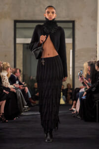 Model walks a runway in a black cropped top, dramatic ruffled neckpiece, and layered pleated skirt; audience sits along the catwalk.