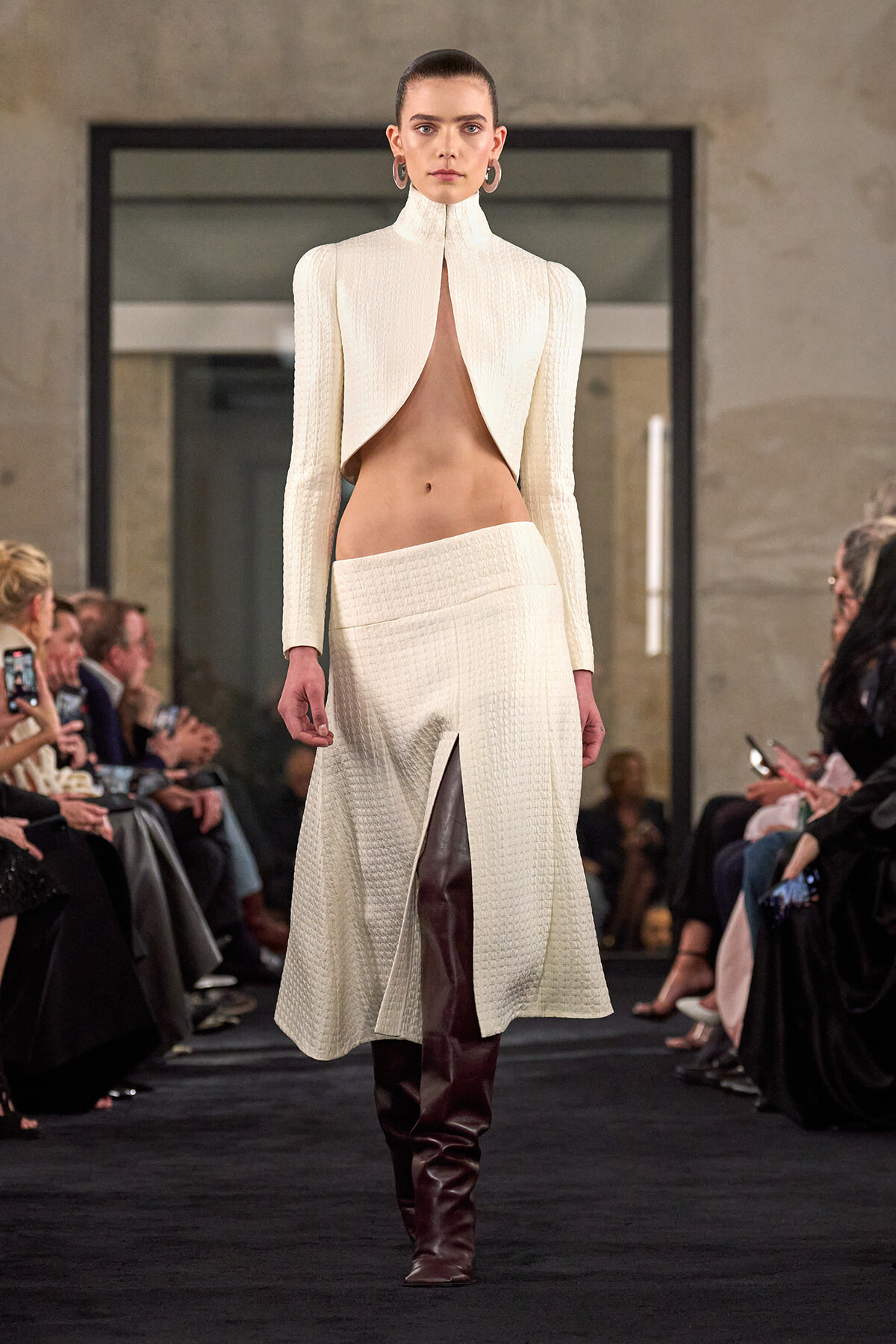 Model on a runway wearing an ivory textured two-piece with a high-neck cape opening at the midriff and burgundy knee-high boots.