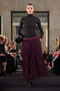 Model walks the runway in a black turtleneck bodysuit and a long purple fringe skirt, carrying a black clutch with oversized gold hoops audience in the background, bold look.