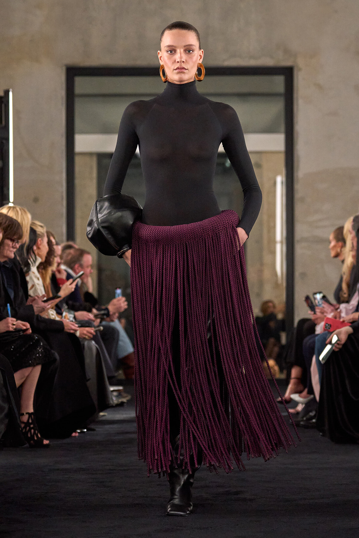 Model walks the runway in a black turtleneck bodysuit and a long purple fringe skirt, carrying a black clutch with oversized gold hoops audience in the background, bold look.