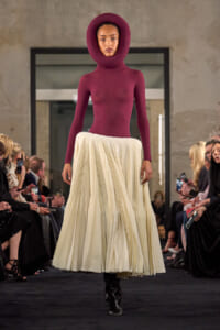 Model walks the runway in a burgundy hooded bodysuit paired with a cream pleated midi skirt, audience visible on both sides.