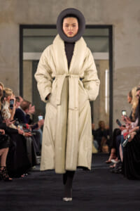 Model walks a beige puffer coat with belt and oversized round black neck collar on a runway.
