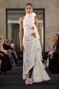 Model walks a runway in a white asymmetric gown with cutouts, tied scarf at the neck, and draped skirt details.