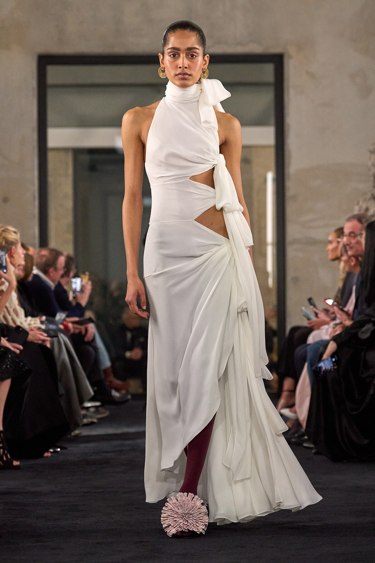 Model walks a runway in a white asymmetric gown with cutouts, tied scarf at the neck, and draped skirt details.