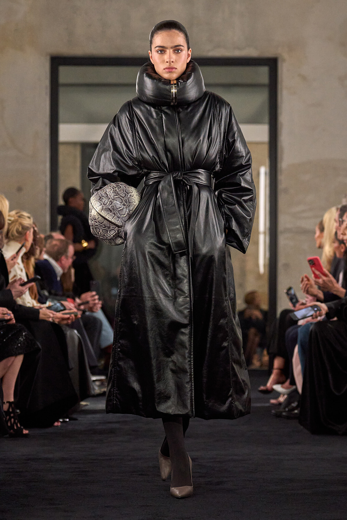 Model walks a runway in a glossy black belted puffer coat, holding a textured ball-shaped clutch; audience seated along the sides.