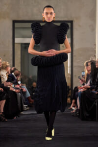 Model walks the runway in a black high-neck dress with oversized ruffled sleeves and a layered ruffle skirt, gold hoop earrings.