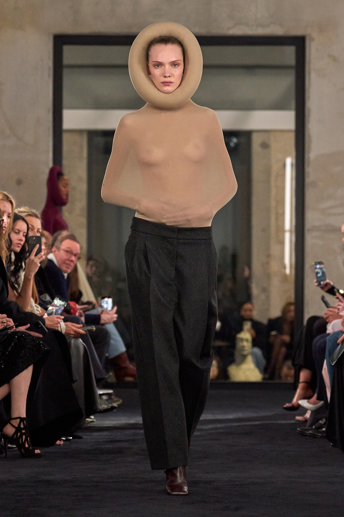 Runway model in a nude-tone sculptural top with a large circular neck collar, black trousers, and ankle boots; spectators line the catwalk.