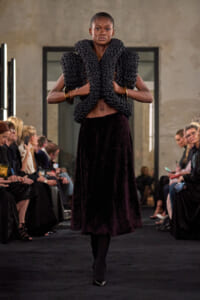 Model walks a fashion show wearing a sculptural black chain-wrapped top over a dark velvet skirt; audience seated on both sides watch the runway.