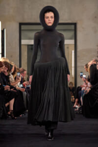 Model walks the runway in a black sculptural outfit: a form-fitting bodysuit with a large circular neckpiece and a long pleated skirt, audience on both sides.