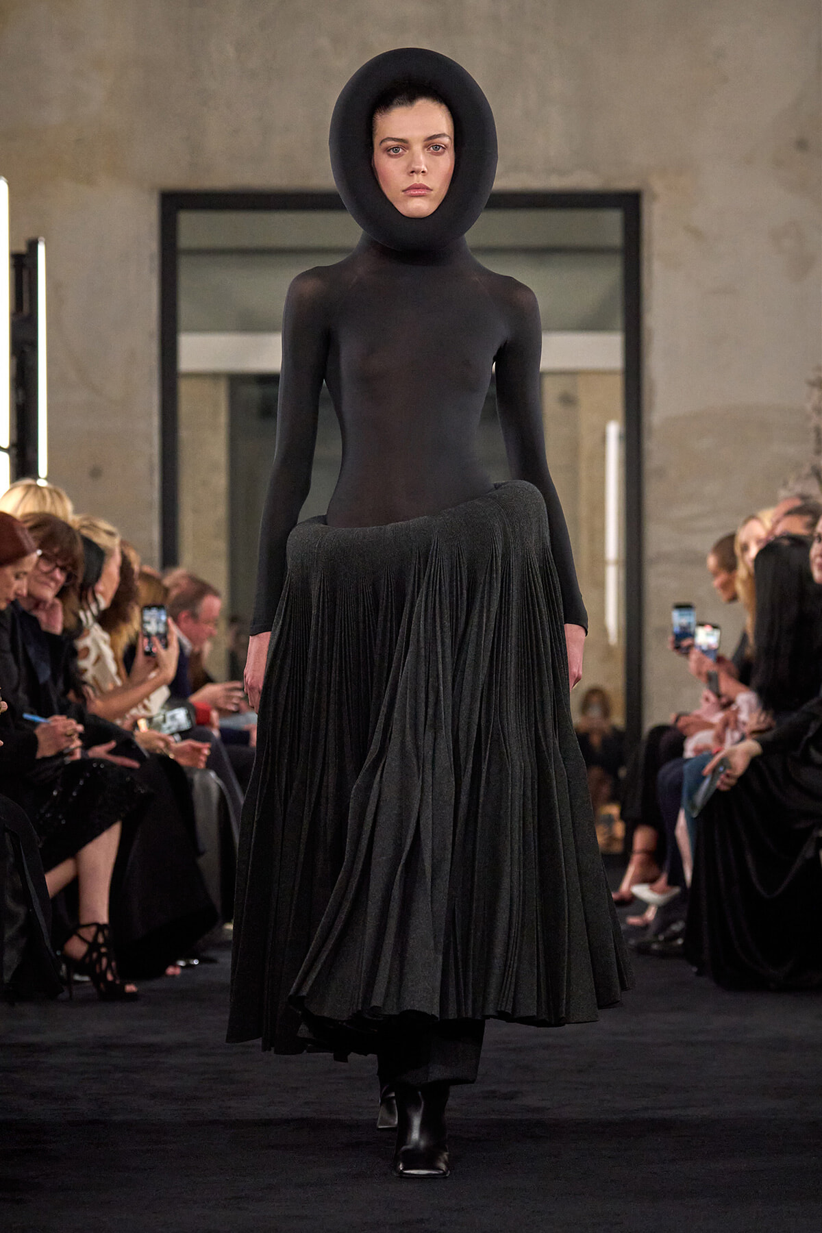 Model walks the runway in a black sculptural outfit: a form-fitting bodysuit with a large circular neckpiece and a long pleated skirt, audience on both sides.