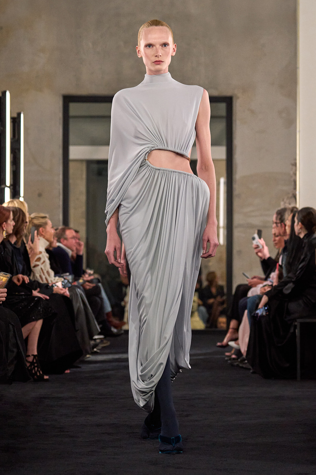 Model walks the runway in a pale gray draped gown with a twisted side cut-out at the waist and flowing gathered skirt.