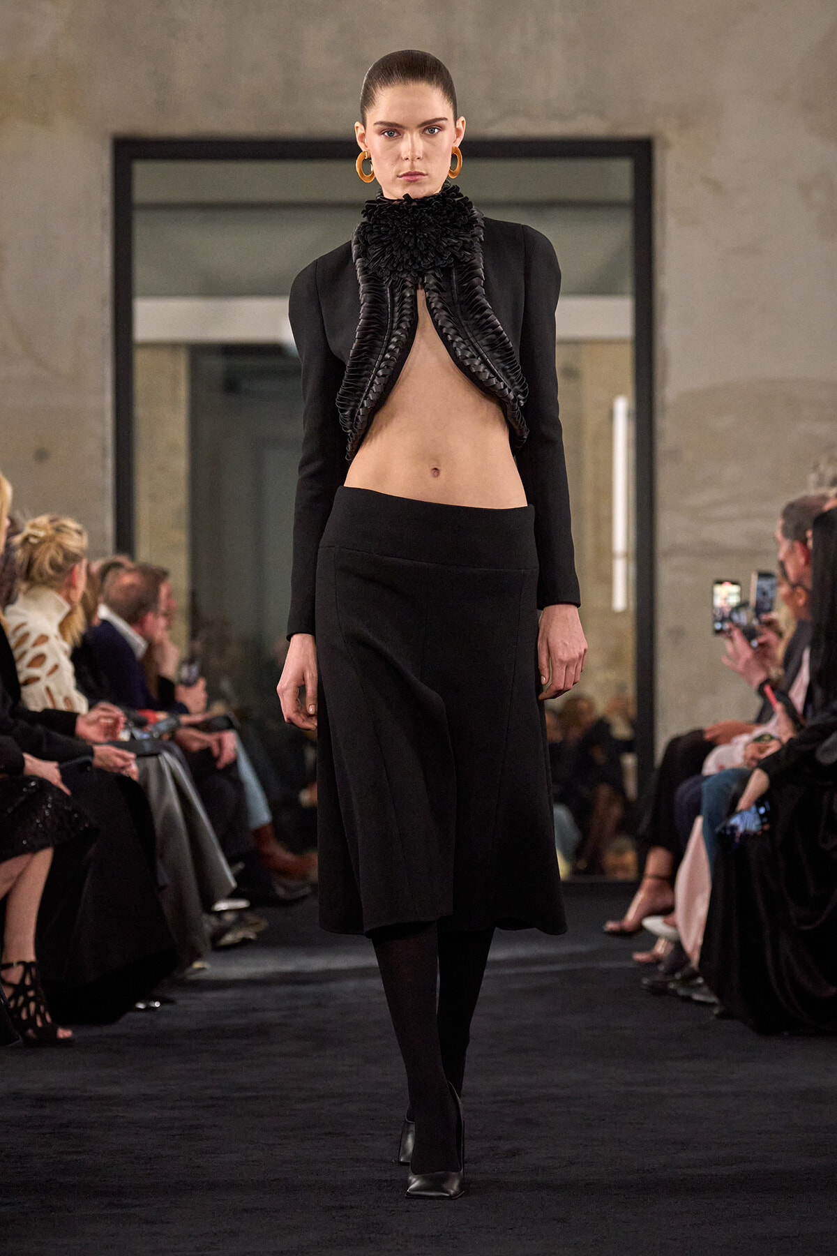 Model walking a fashion runway in a black ensemble with a dramatic sculpted ruffled collar, exposing midriff. A sleek midi skirt and heels complete the look.