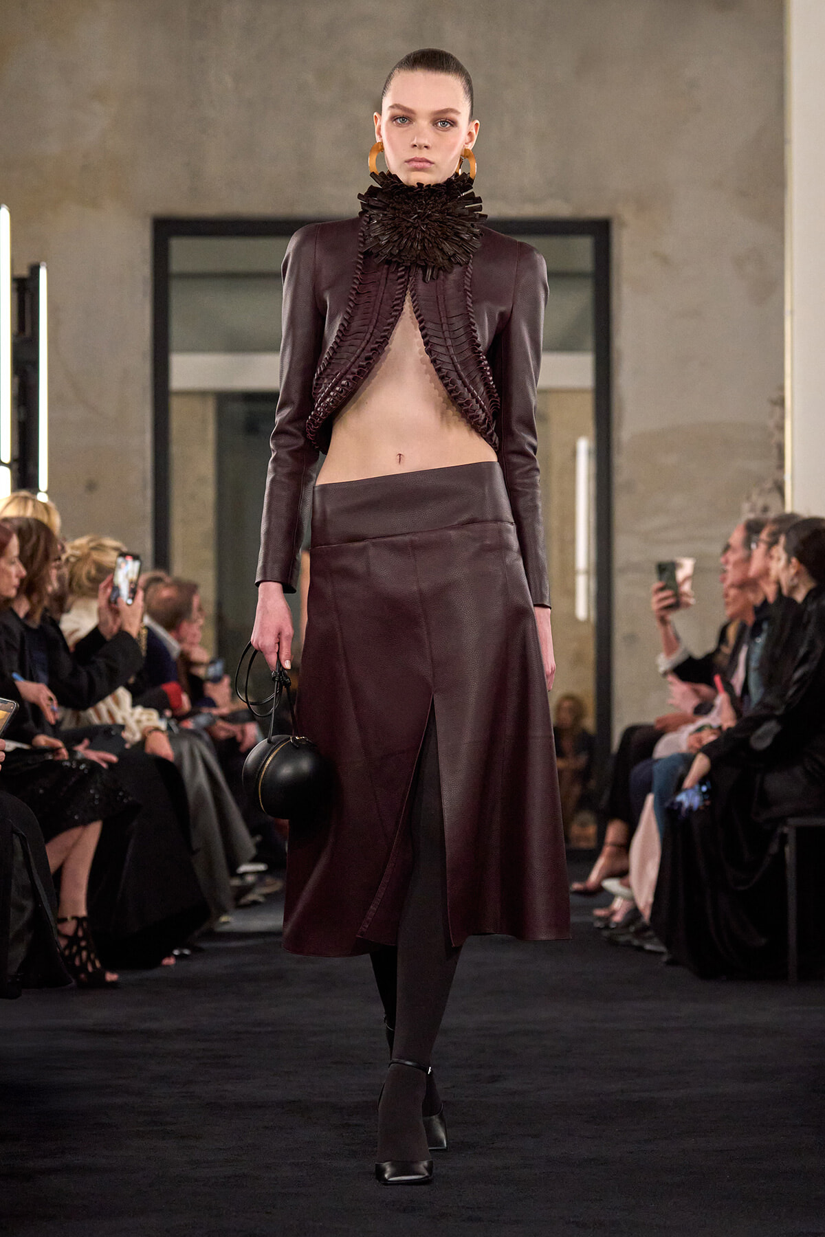 Model walks the runway in a burgundy leather ensemble: cropped jacket with a sculpted ruffled high collar, matching mid-length skirt with a front slit, black tights, and ankle-strap heels, holding a small round black bag.