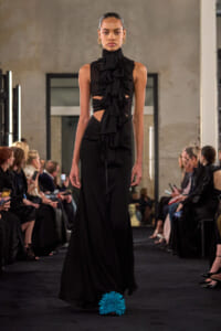 Model walks a runway in a long black gown with dramatic ruffles at the chest and waist; gold hoop earrings and a sleek updo.