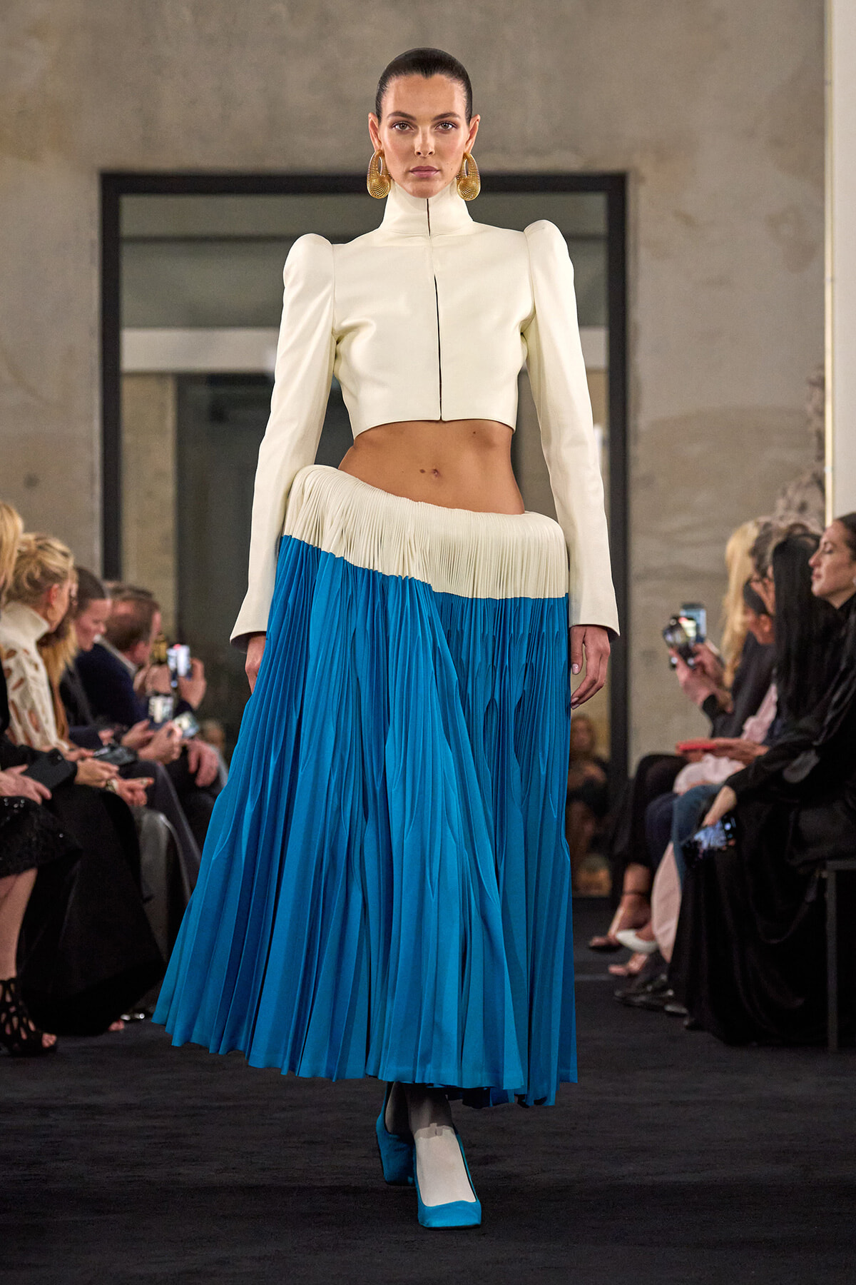 Model on a runway wearing an ivory cropped jacket with a high collar and puffed shoulders paired with a blue pleated maxi skirt.