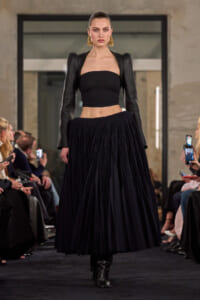 Model walks the runway in a black bandeau top, a full pleated midi skirt, and sculpted leather puff sleeves with oversized gold hoop earrings.