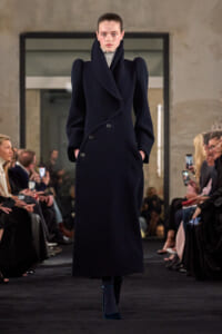 Model walking a runway in a navy, oversized double-breasted coat with a high, sculptural collar.