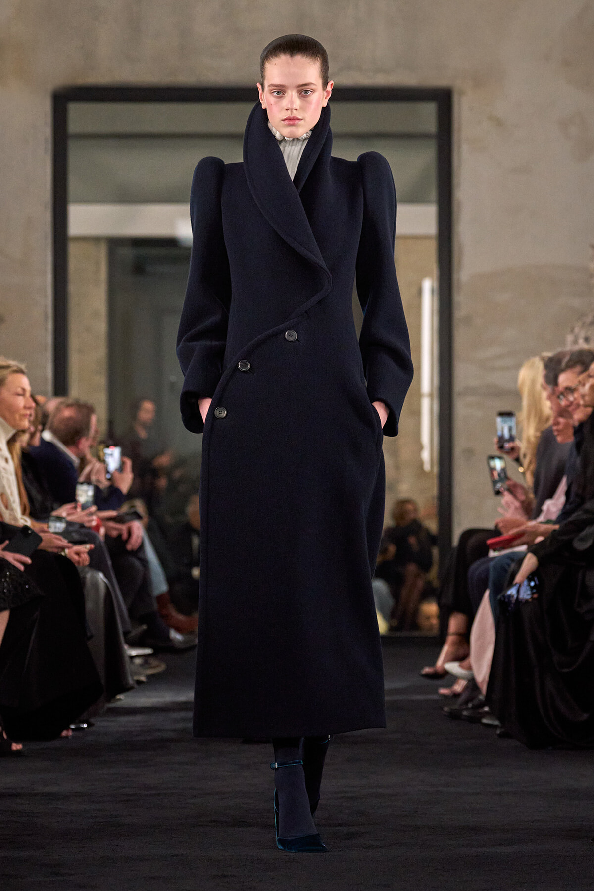 Model walking a runway in a navy, oversized double-breasted coat with a high, sculptural collar.