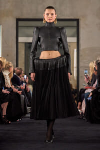Model walks the runway in a black cropped leather jacket with a high neck paired with a long pleated black skirt, gold hoop earrings visible.