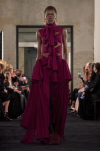 Model walking a runway in a burgundy cutout gown with a large bow at the neck and dramatic draped skirt.